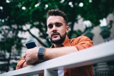 Aşağıdaki hippi görünümlü sakallı sıradan turuncu tişörtlü adamdan. Elinde akıllı bir telefon, çitlere yaslanmış, caddede duruyor ve kameraya bakıyor.