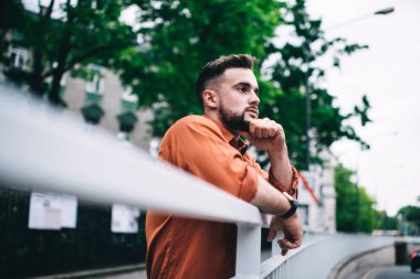 Sakallı bir adamın metal korkuluklara yaslanıp şehrin bulanık arka planını düşünürken yüzünü çevirmesi.