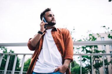 Ciddi sakallı, turuncu tişörtlü, el ele tutuşan ve şehir caddesinde parmaklıkların yanında akıllı telefondan konuşurken yüzünü çeviren adam.