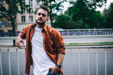 Kendine güvenen sakallı adam el ele tutuşup kameraya bakarken sokağın yakınındaki metal çitlere yaslanıyor.