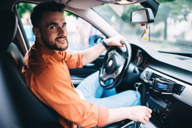 Pahalı bir arabada oturan ve arka plandaki bulanık kameraya bakarken direksiyon sallayan sıradan giyinen odaklanmış sakallı erkek sürücünün yan görüntüsü.