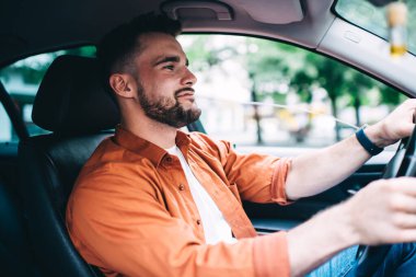 Turuncu tişörtlü, sakallı, gülümseyen ve direksiyon sallayan, şehrin bulanık arka planında...