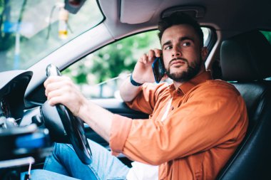 Pahalı bir arabada oturan, direksiyonu çeviren, akıllı telefondan konuşan ve arka planda bulanık duran erkek sürücünün yan görüntüsü.