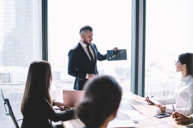 Modern ofis binasının penceresinin yanında dururken şık saç kesimi olan sakallı bir yönetici yönetim kuruluna tabloyu sunuyor.
