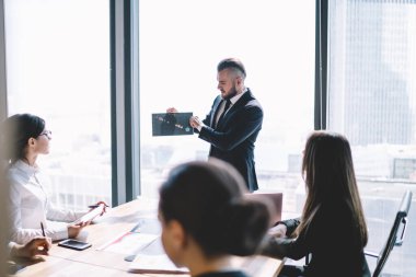 Ciddi sakallı, resmi giyinmiş, şık saç kesimi olan ciddi bir iş adamı modern ofis binasının penceresinin yanında dururken yönetim kuruluna tabloyu sunuyor.