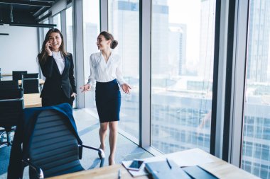 Neşeli etnik kadın yönetici iş ortağıyla cep telefonunda konuşuyor. Ofis binasının yakınında yürüyen gülümseyen kadın iş arkadaşıyla. 