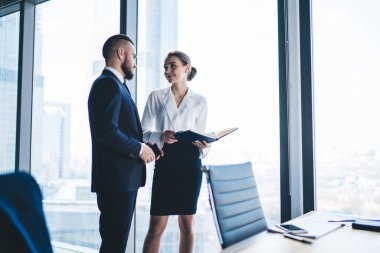 Resmi kıyafetleri içinde tanınmayan bir erkek CEO, modern ofis binasının penceresinde dururken elinde not defteriyle kadın muhasebeciyle konuşuyor.