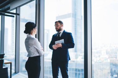 Kadın yönetici, şehir manzaralı modern ofis binasının penceresinin yanında dururken erkek iş ortağıyla konuşuyor.