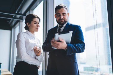Takım elbiseli erkek yönetici, modern ofis odasının penceresinin yanında dururken tabletten kadın asistana resmi giysiler içinde bilgi gösteriyor.