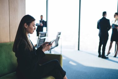 Zarif takım elbiseli ciddi etnik kadın akıllı telefonuyla mesajlaşırken ofisin bulanık arka planında rahat bir koltukta oturuyor.