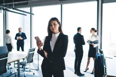 Ciddi Asyalı patron akıllı telefon kullanarak modern ofisin bulanık arka planında dururken kameraya bakıyor.