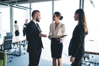 Sakallı adam, modern ışık ofisinde bayan iş arkadaşının yanında durup açık defterli bir kadınla konuşuyor.
