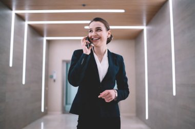 Neşeli ve neşeli bir ofis çalışanı. Parlak bir koridorda durmuş, akıllı telefondan konuşurken gülümsüyor ve gözlerini kaçırıyor.