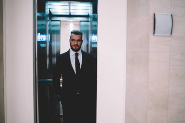 Koyu renk saçlı, stil sahibi, ciddi düşünceli, zarif elbiseli bir adam asansör kapıları çağdaş ofiste kapanıyorken dikiliyor ve başka tarafa bakıyor.