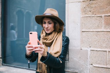 Deve rengi şapkalı, siyah ceketli, modern akıllı telefonlu ve eski mimari binanın yanında dururken selfie çeken neşeli, şık bir kadın.