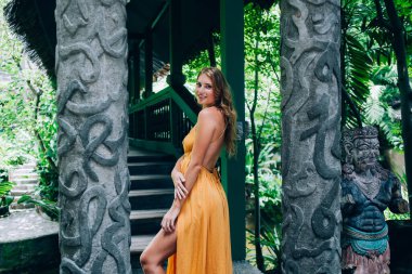 Uzun sarı saçlı, sırtı açık turuncu maksi elbiseli ve Bali tropikal ormanlarında köprüde ayakta duran harika gülümseyen genç bir kadın.