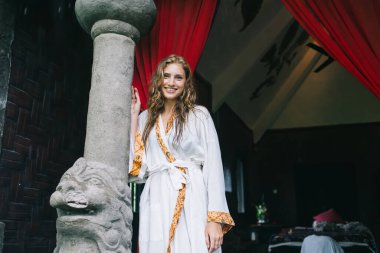 Beyaz cüppeli, dalgalı saçlı, Budist aslanla taş sütunun yanında kameraya bakarken gülümseyen genç bayan.