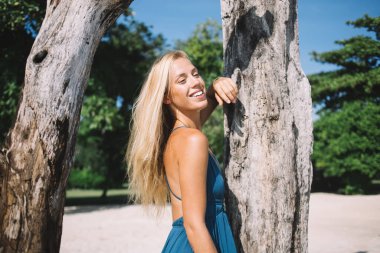 Sarışın, mutlu, dişlek, ağaçlara dokunan ve güneşli bir günde kumsalda kahkahalar atan genç bir kadın.