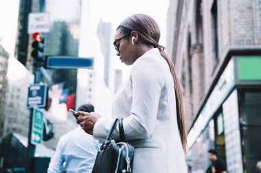 Kablosuz kulaklıklı, müzik dinleyen ve akıllı telefondan mesajlaşan meşgul Afro-Amerikan kadının düşük açısı. Şehir sokaklarının bulanık arka planında yürürken.