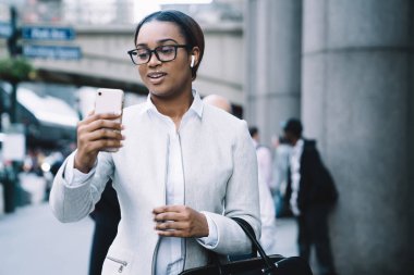 Beyaz takım elbiseli, kablosuz kulaklıklı, sokakta yürürken cep telefonuyla konuşan akıllı, Afro-Amerikalı bir kadın. 