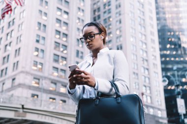 Gözlüklü başarılı Afrikalı Amerikalı kadın girişimci arka planda gökdelenler ve ABD bayraklarıyla akıllı telefondan mesaj yolluyor.