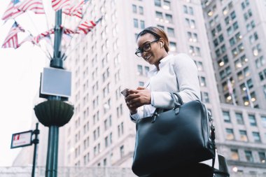 Başarılı gülümseyen Afrikalı Amerikalı yönetici kadın şık takım elbiseli, siyah deri çantalı, akıllı telefondan mesaj atan arka planda gökdelenlerle şehir merkezinde duran 