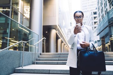 Siyah iyi giyimli, kulaklıklı ve çantalı ciddi bir bayan şehir merkezindeki ofis binaları arasında işe giderken cep telefonunu kontrol ediyor.