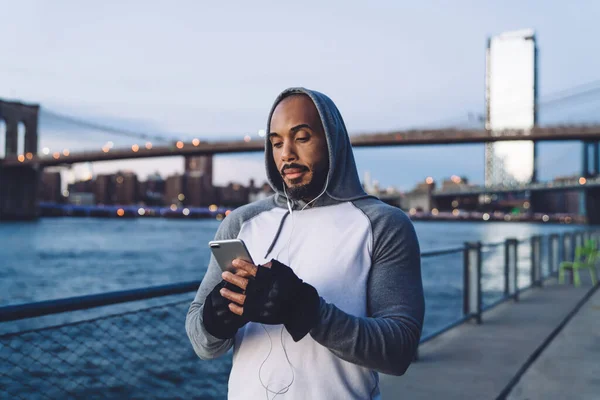 年轻的非裔美国人站在耳机里听音乐 一边在河边和桥上用智能手机发短信 — 图库照片