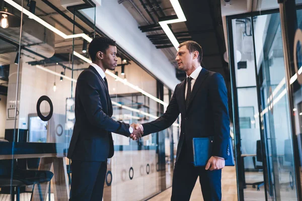 Multiethnic busy entrepreneurs in lounge suits concluding an agreement while meeting at modern office hall with glasses walls and shaking hands