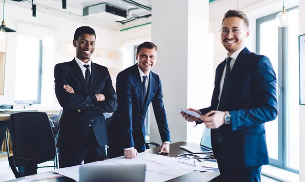 Positive smiling diverse businessmen in suits standing around table with papers and scheme of project at office looking at camera
