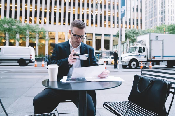 严肃而专注的商人 身穿黑色办公室套装 戴着眼镜 坐在街头咖啡馆里 在模糊的背景下 用电话对文档报告拍照 — 图库照片
