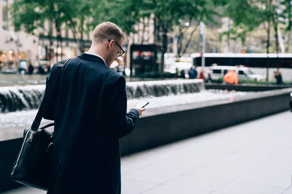 眼鏡と革のブリーフケースが立っている黒いコートの従業員のバックビューと路上のぼやけた背景にスマートフォンを使用して — ストック写真