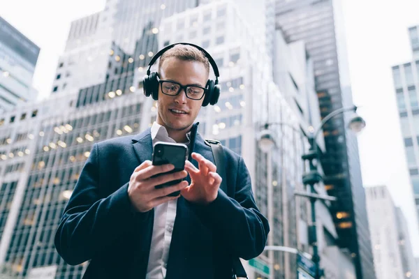 Gözlüklü, resmi ve yakışıklı bir erkek çalışandan kablosuz kulaklıkla müzik dinlerken ticari binaların bulanık arka planında cep telefonuna göz gezdiriliyor. 