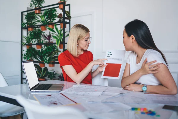 Concentrated young lady in glasses pointing at document creating collaborative startup with ethnic woman in casual wear at modern work space