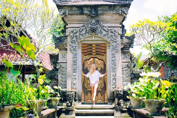 Full body happy woman smiling and swinging leg while standing near door of aged oriental shrine on sunny day in tropical garden