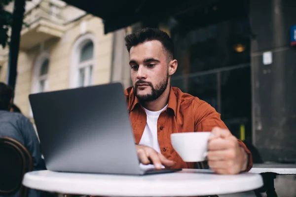 情報を求めてコーヒーとサーフィンノートパソコンと街の通りでクールなカフェテリアに座っている若いカジュアルな男を驚かせた — ストック写真