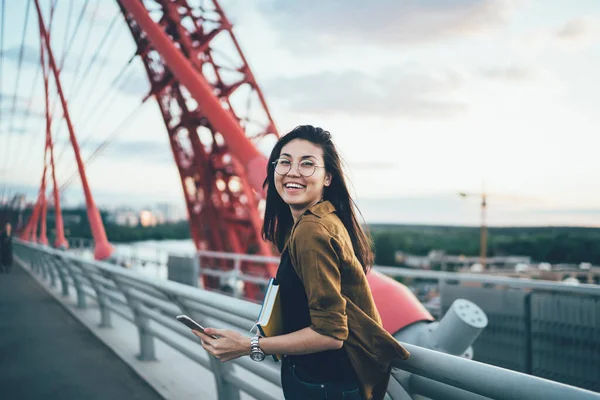 Portrait of cheerful asian international student in eyewear spending time outdoors on bridge holding mobile phone for blogging, smiling female traveler satisfied with roaming for search destination