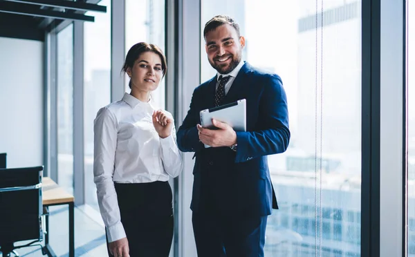 Pretty young woman in formal wear and bearded man with tablet smiling and looking at camera while standing near window in modern office together