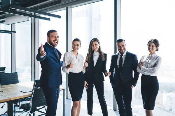 Diverse successful office workers in formal wear cheerfully smiling and gesticulating for camera while standing against huge bright window together