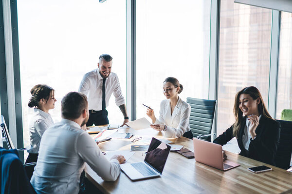 Группа бизнес-работников, сидящих за столом на встрече в светлом офисе и глядящих на лидера команды стоя и говоря о процессе работы со смехом
 