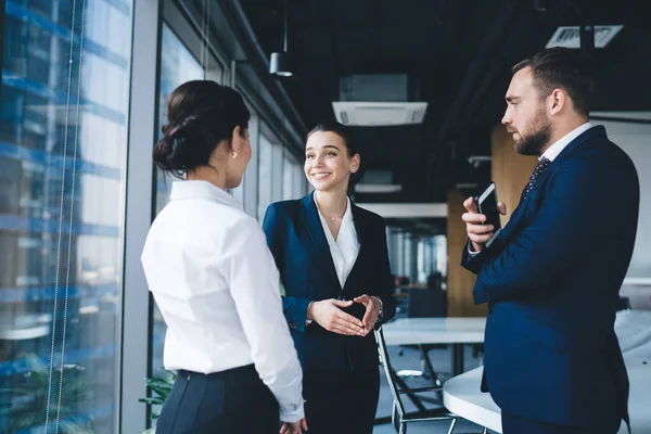 Friendly young woman in formal wear smiling and talking with colleagues while standing in contemporary office and talking during break