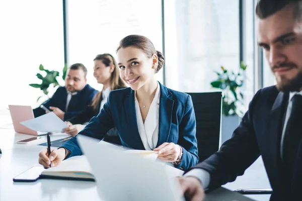 Smart female analyst working with documents writing in planner and looking at camera with charming smile sitting at table on meeting