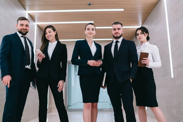 現代の多様なよく服を着た男性と女性の企業の労働者のグループガジェットやプランナーが現代のオフィス廊下に一緒に立っている — ストック写真