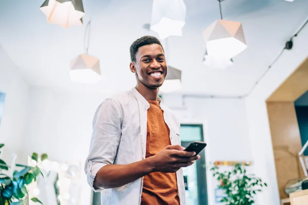 Half length portrait of cheerful hipster guy with black dark skin smiling at camera during leisure time for smartphone wireless networking, happy African American man holding digital mobile technology