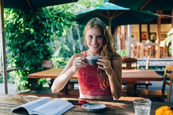 Happy adult female in red shirt looking at camera and holding cup with beverage while sitting at wooden table with book and smartphone on cafe terrace under big turquoise umbrella at Bali resort in fair weather