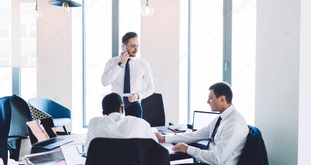 Thoughtful adult making phone call boss in glasses and elegant clothes standing and waiting results while working with multiracial colleagues in light modern office