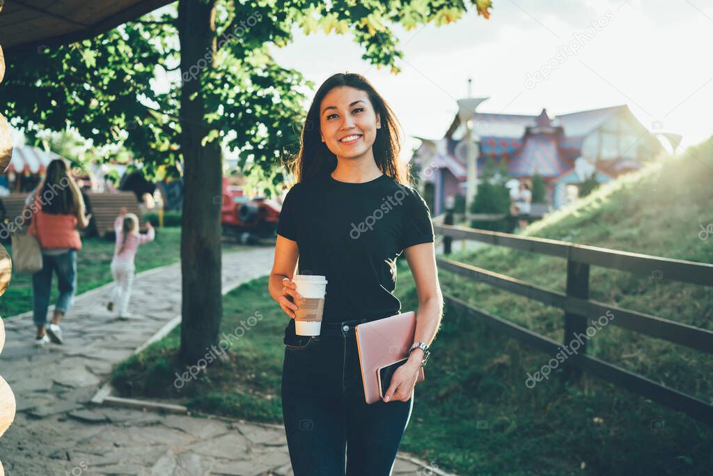 Portrait of cheerful asian hipster girl in casual wear standing in city park with coffee to go cup enjoying weekends, smiling good looking female student having break outdoors resting on evening 