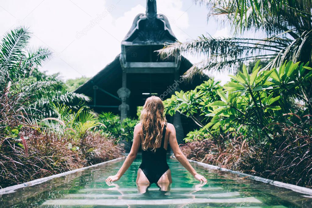 Back view of adult lady in black swimsuit going along turquoise narrow swimming pool with crystal clear water in garden with green and brown exotic plants against recreation area during hot sunny day