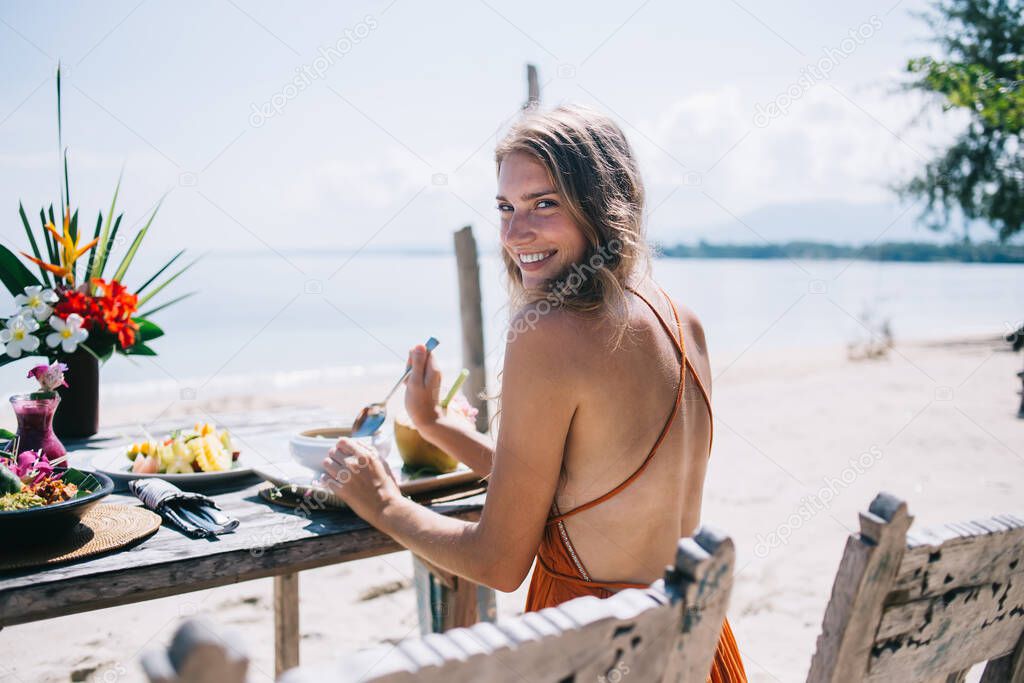Back view of cheerful female person on vacation having breakfast at beautifully served table and looking over shoulder at camera on seashore