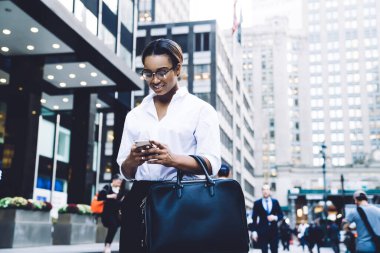 Büyük siyah çantalı şık Afro-Amerikan iş kadını akıllı telefondan iyi haberler okuyor ve modern ofis binalarının yanında yürürken gülümsüyor.